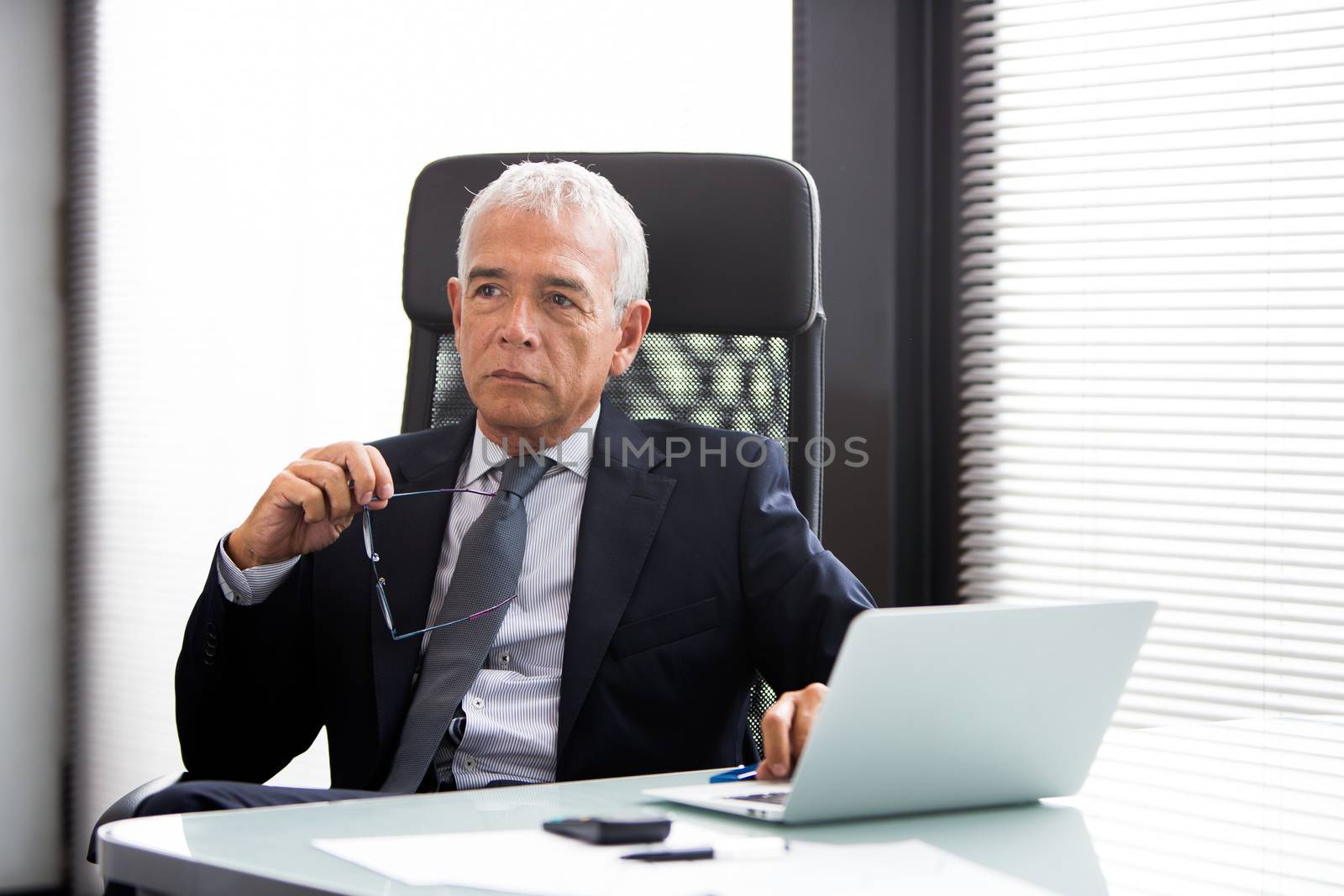 Half length portrait of a thoughtful businessman in the office with laptop computer and holding glasses 
