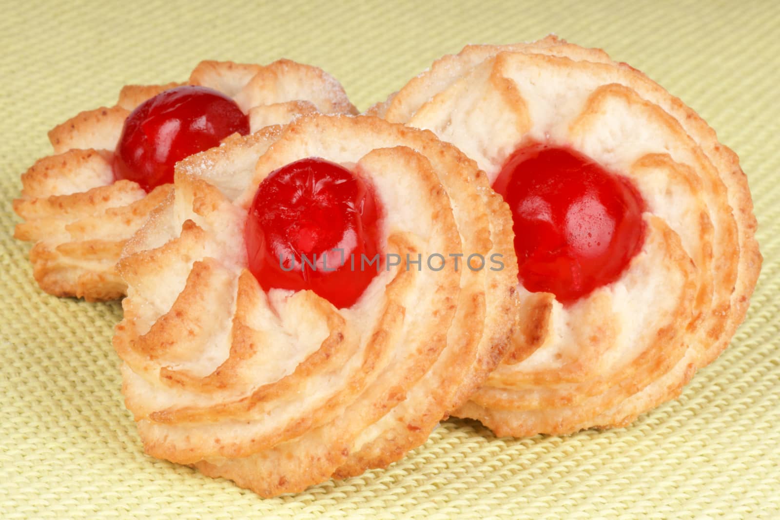 Three almond pastries decorated with red candied cherries over a light green fabric background
