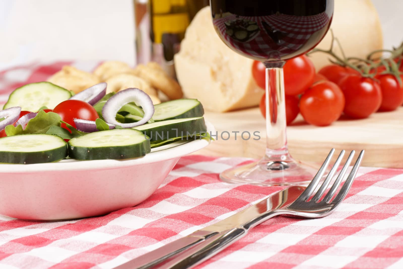 Close-up of a prepared table with an healthy salad, a glass of red wine, fork and knife. In the background some red tomatoes and parmesan cheese. Selective focus.