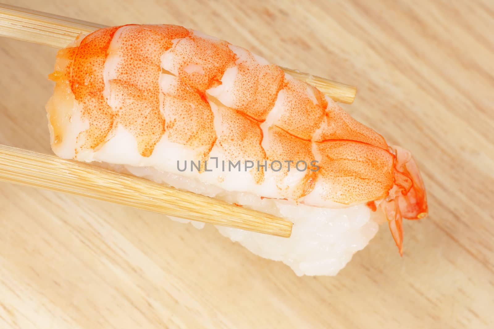 Wooden chopsticks holding a piece of sushi with shrimp, over a wooden background