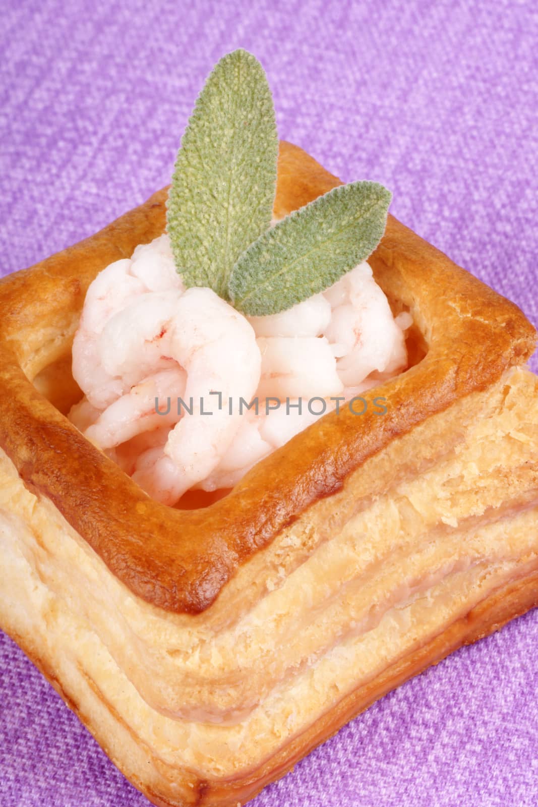 Close-up of a squared vol-au-vent stuffed with small boiled shrimps decorated with sage leaves over a purple background