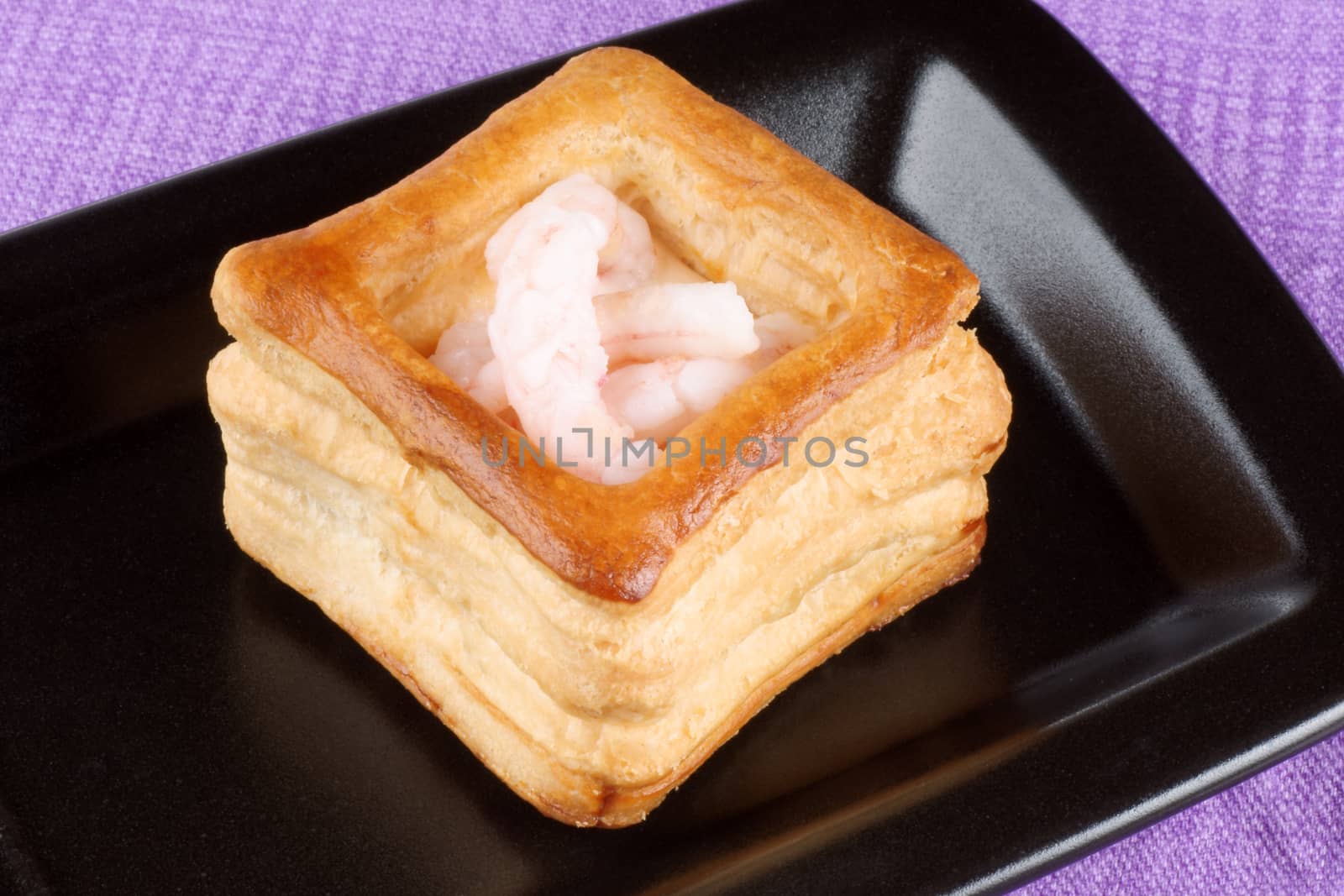 Close-up of a squared vol-au-vent stuffed with small boiled shrimps on a black plate