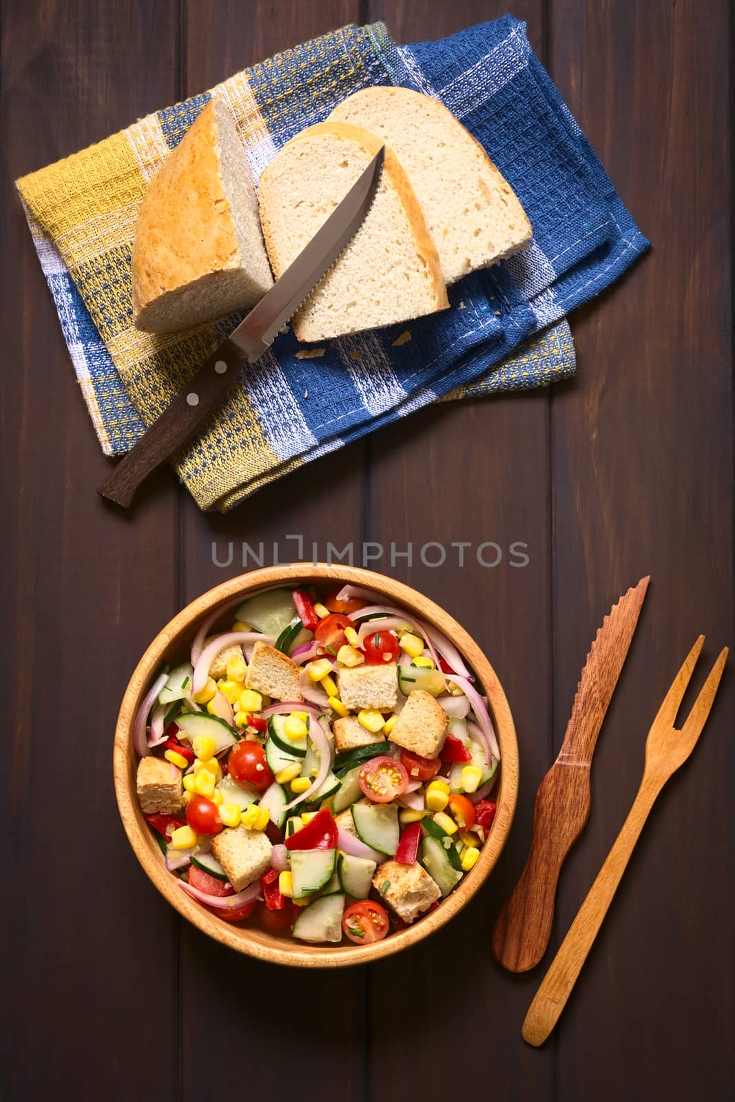 Overhead shot of fresh vegetable salad made of sweet corn, cherry tomato, cucumber, red onion, red pepper, chives with croutons in wooden bowl, photographed on dark wood with natural light