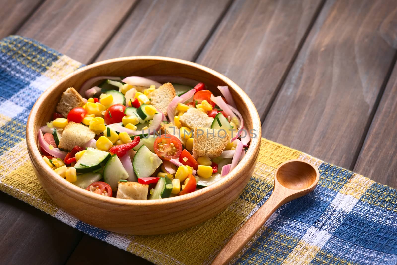 Fresh vegetable salad made of sweet corn, cherry tomato, cucumber, red onion, red pepper, chives with croutons in wooden bowl, photographed on dark wood with natural light (Selective Focus, Focus one third into the salad)