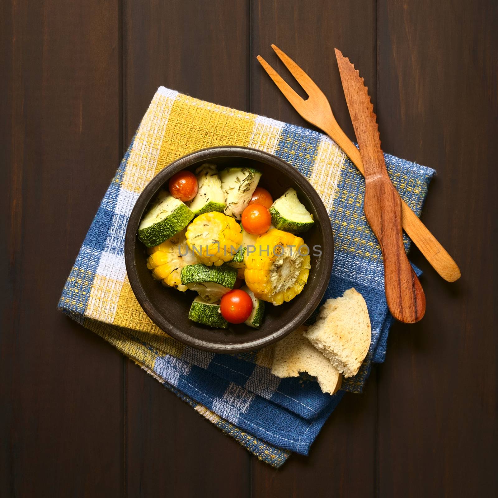 Overhead shot of a bowl of baked vegetables of sweet corn, zucchini, cherry tomato with thyme, toasted bread slice and wooden cutlery on the side, photographed on dark wood with natural light