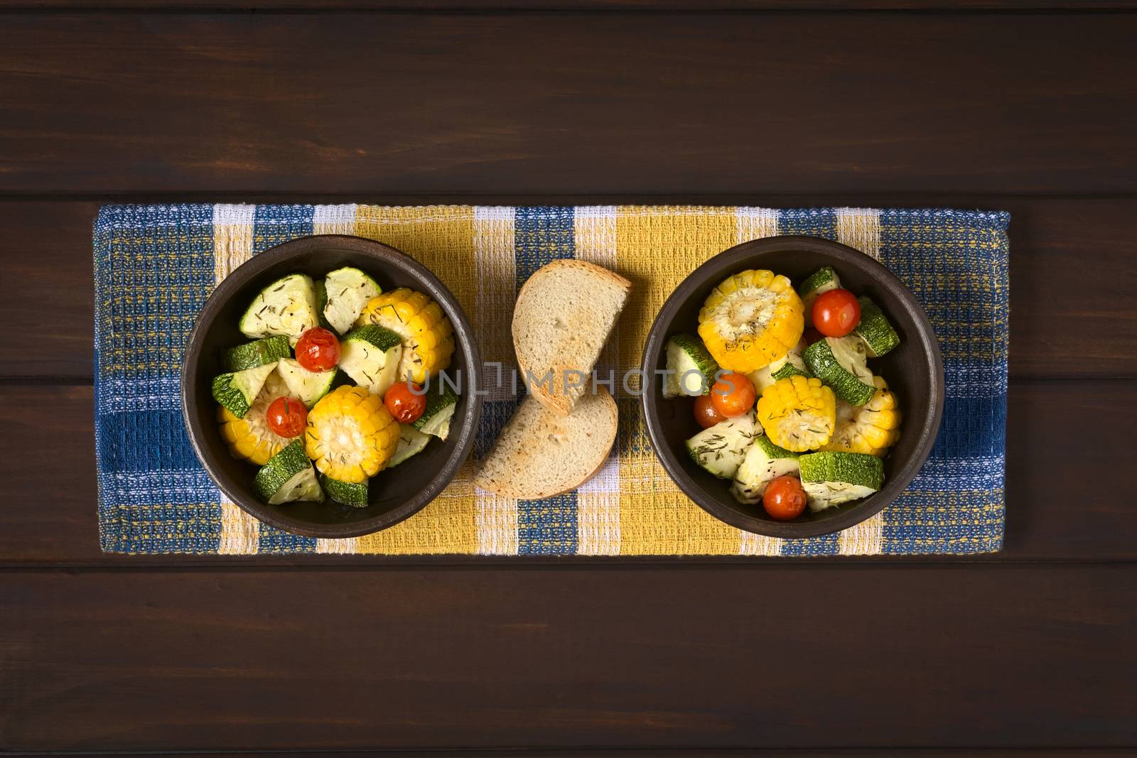 Overhead shot of two bowls of baked vegetables of sweet corn, zucchini, cherry tomato with thyme, toasted bread slices in the middle, photographed on dark wood with natural light