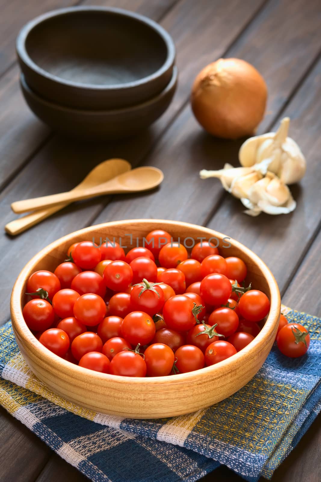 Cherry tomatoes in wooden bowl with garlic, onion, bowls and wooden spoons in the back, photographed on wood with natural light (Selective Focus, Focus one third into the tomatoes)