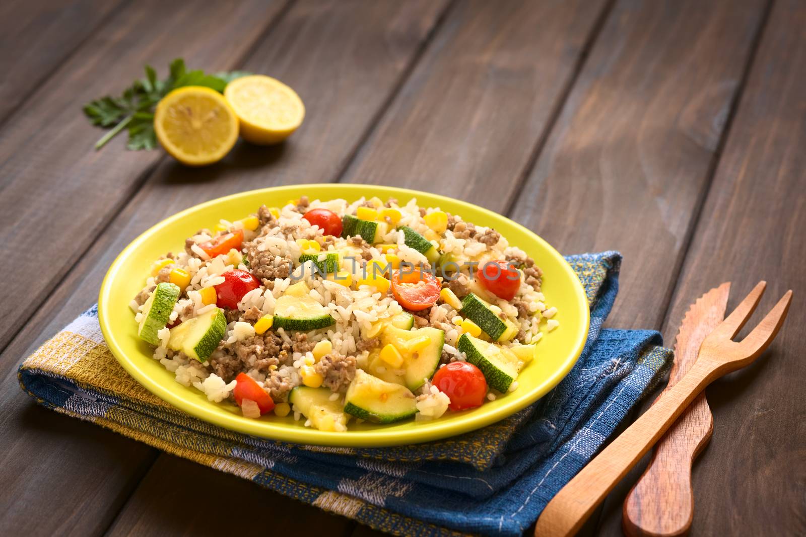 Rice dish with mincemeat and vegetables (sweet corn, cherry tomato, zucchini, onion) served on plate with wooden cutlery on the side, photographed on dark wood with natural light (Selective Focus, Focus in the middle of the dish)