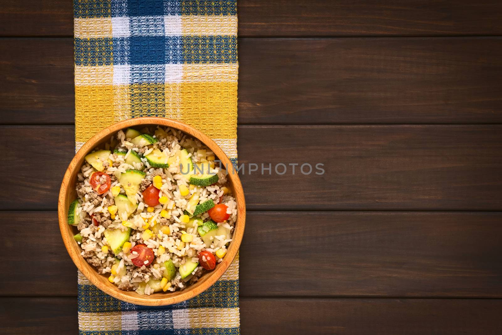 Overhead shot of rice dish with mincemeat and vegetables (sweet corn, cherry tomato, zucchini, onion) in wooden bowl, photographed on dish towel on dark wood with natural light