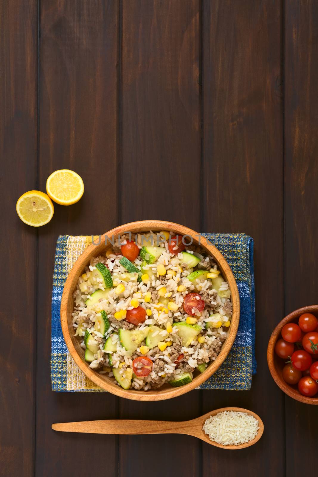Overhead shot of rice dish with mincemeat and vegetables (sweet corn, cherry tomato, zucchini, onion) in wooden bowl with ingredients on the side, photographed on dish towel on dark wood with natural light