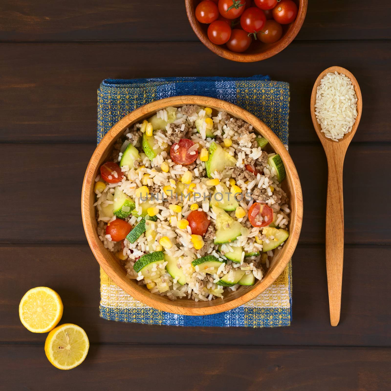 Overhead shot of rice dish with mincemeat and vegetables (sweet corn, cherry tomato, zucchini, onion) in wooden bowl with ingredients on the side, photographed on dish towel on dark wood with natural light