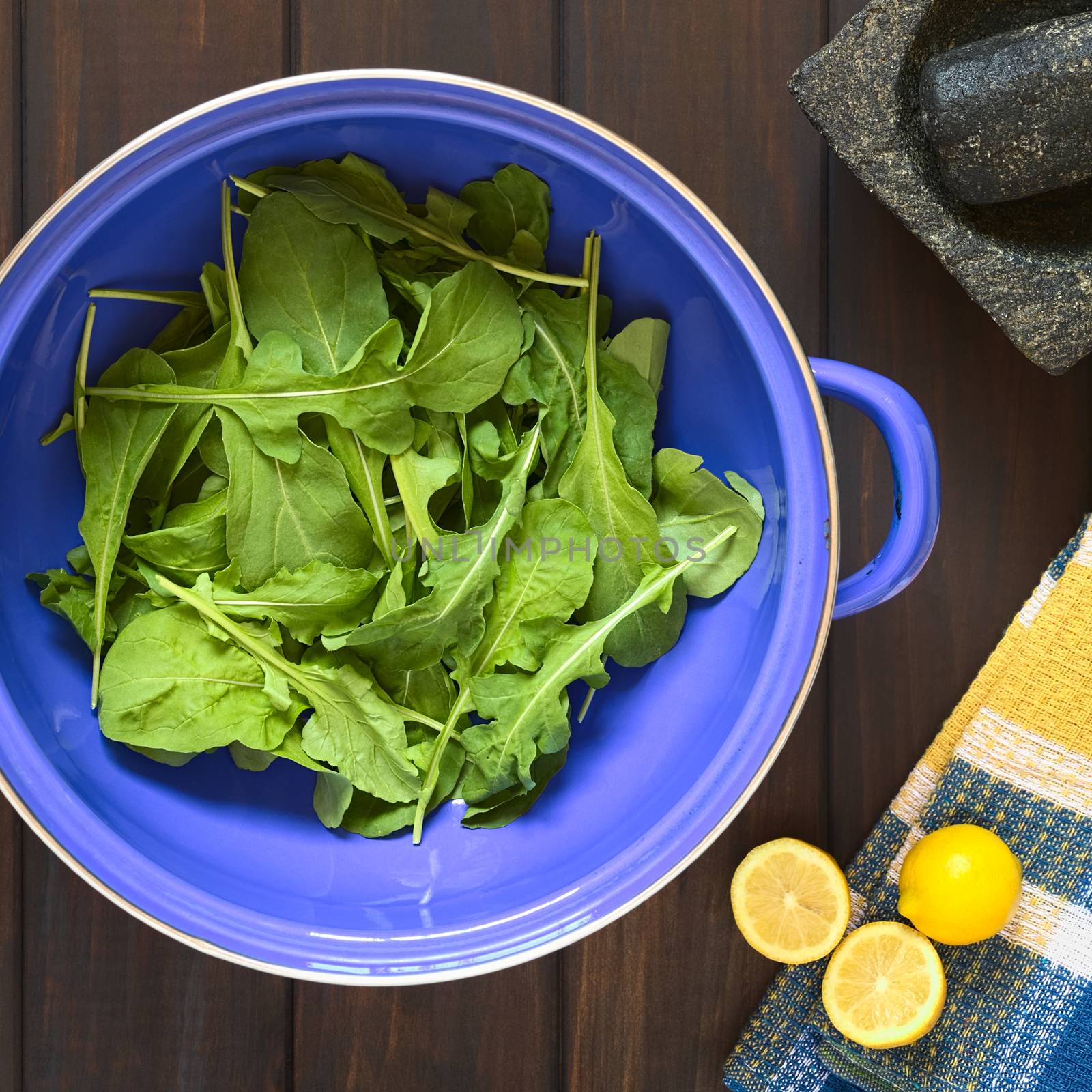 Overhead shot of rucola (lat. Eruca sativa) leaves in blue metal strainer with lemon, mortar and pestle, photographed on dark wood with natural light