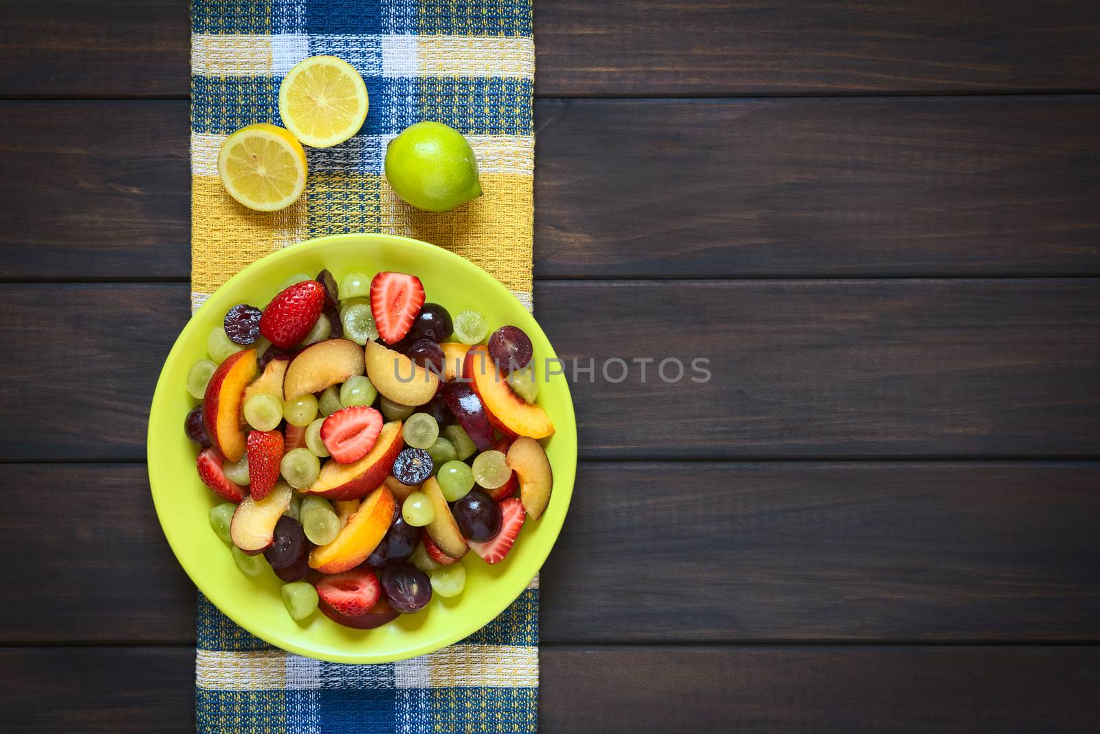 Overhead shot of fresh fruit salad made of grape, strawberry, plum and nectarine served on plate with lemon above, photographed on dark wood with natural light