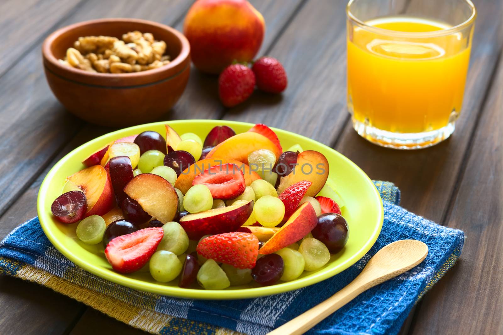 Fresh fruit salad made of grape, strawberry, plum and nectarine served on plate with walnut, nectarine, strawberry, juice in the back, photographed on dark wood with natural light (Selective Focus, Focus in the middle of the salad)