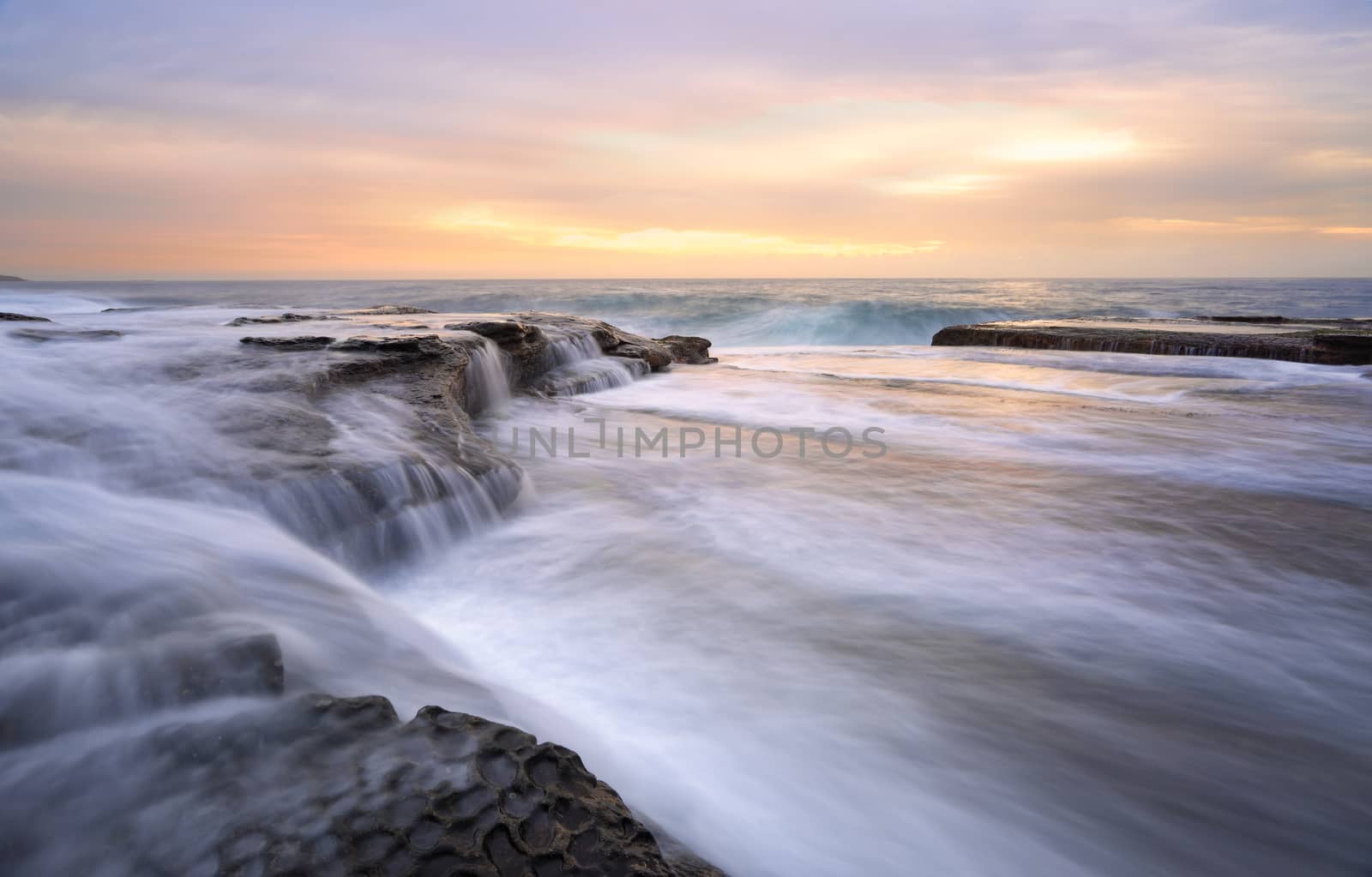 Fast flowing torrents at Curl Curl rock shelf.   One large waves flows off the rocks like a waterfall as the swell builds another wave.   These fast flowing waters feel like whitewater  river flows