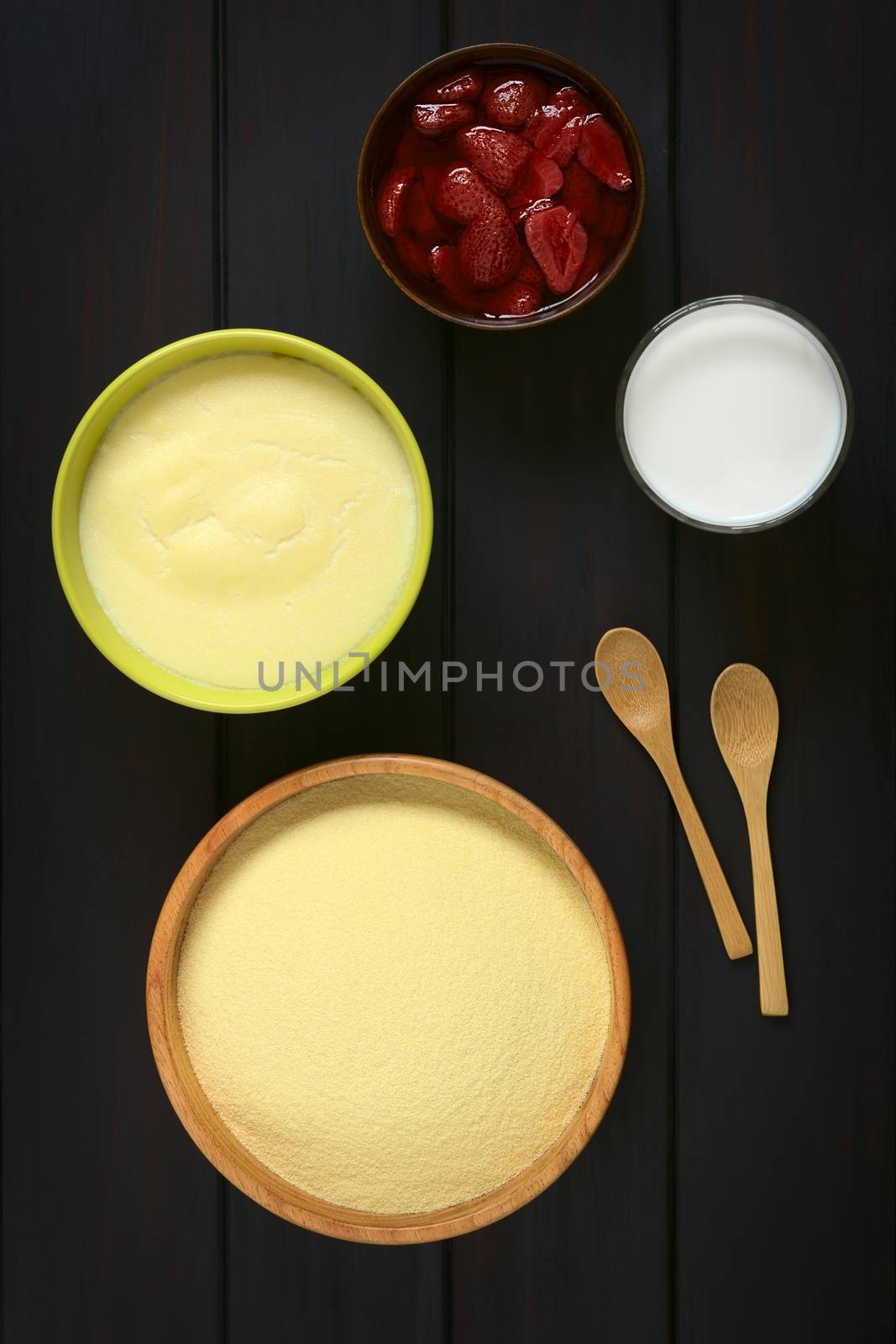 Overhead shot of raw semolina in wooden bowl, with semolina pudding, glass of milk and a bowl of strawberry compote, photographed on dark wood with natural light  