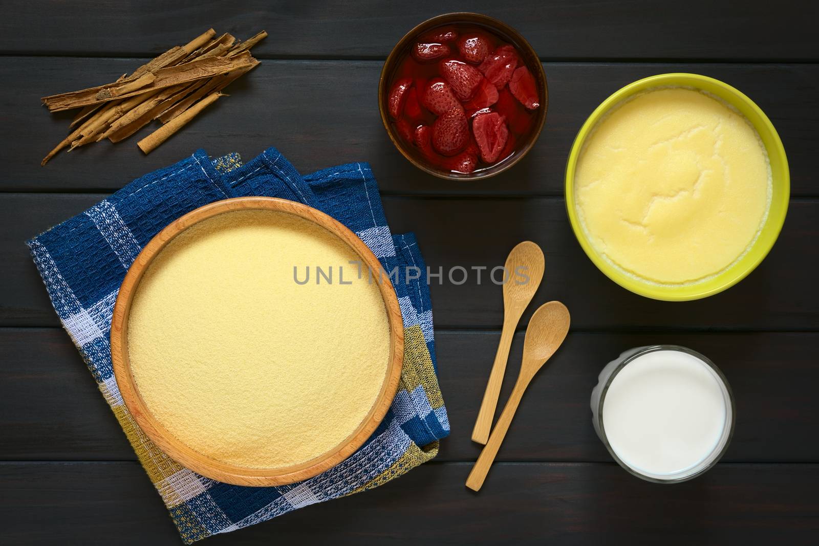 Overhead shot of raw semolina in wooden bowl, with semolina pudding, glass of milk, a bowl of strawberry compote and cinnamon sticks, photographed on dark wood with natural light  