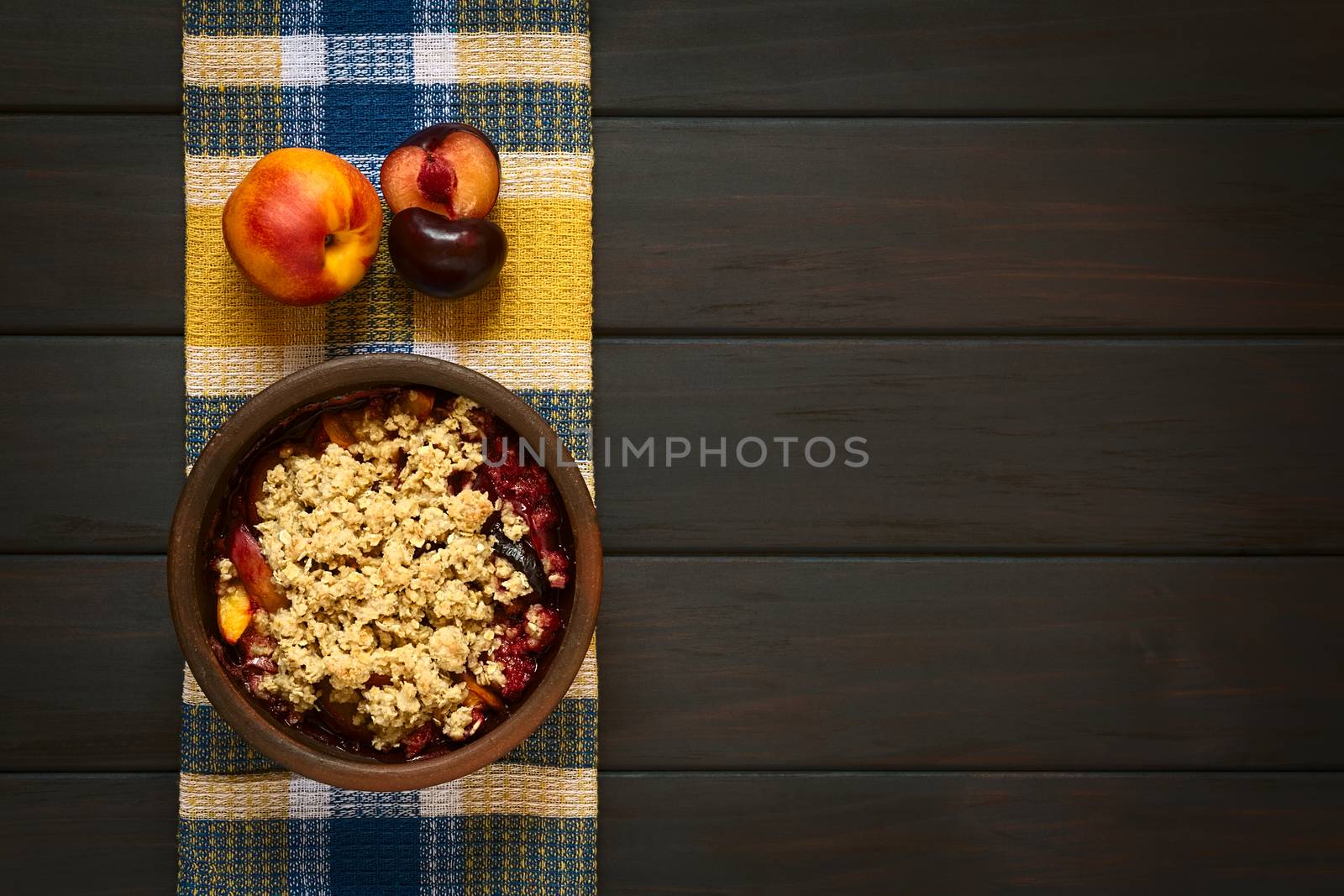 Overhead shot of a rustic bowl filled with baked plum and nectarine crumble or crisp, photographed on cloth on dark wood with natural light