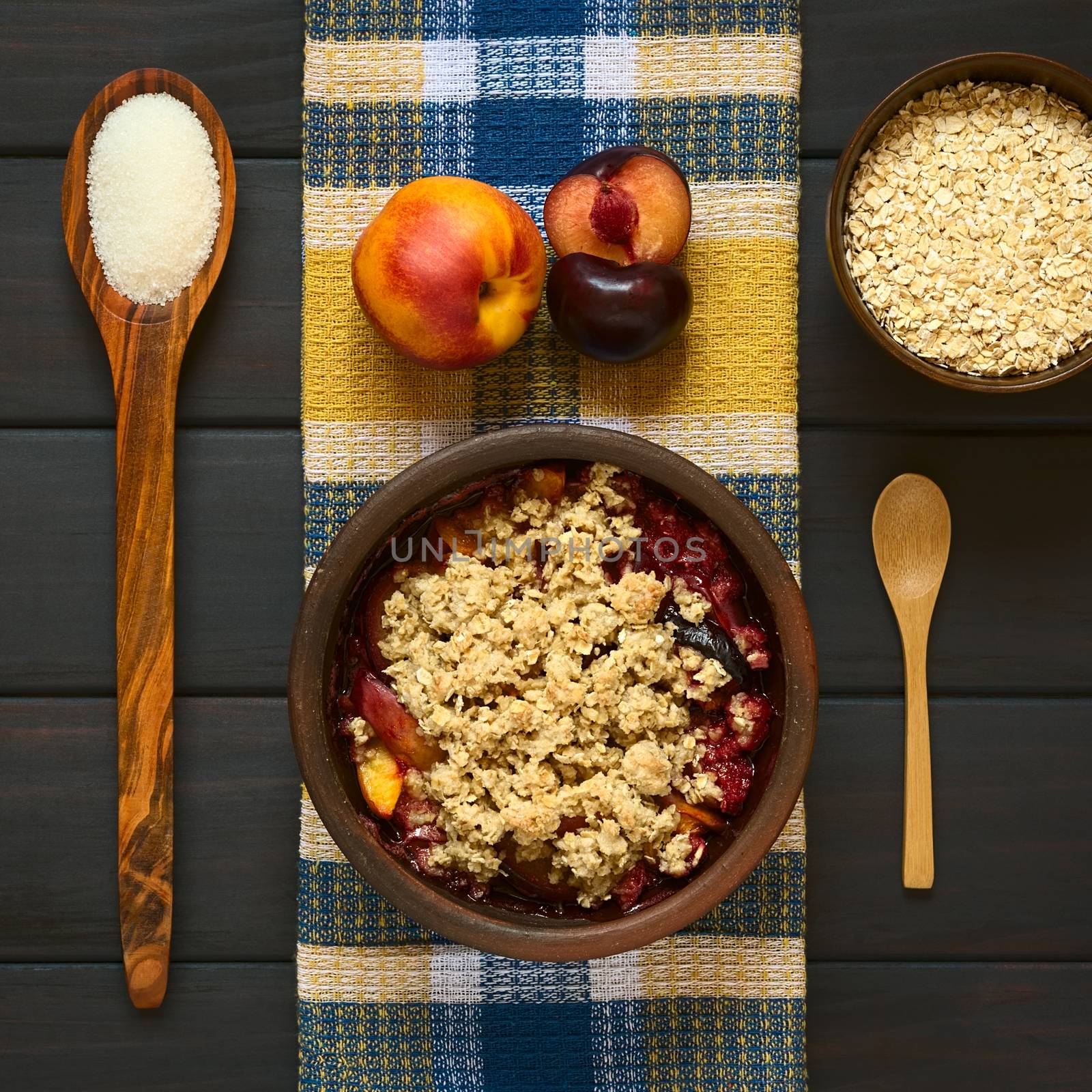 Overhead shot of a rustic bowl filled with baked plum and nectarine crumble or crisp with ingredients on the side, photographed on dark wood with natural light