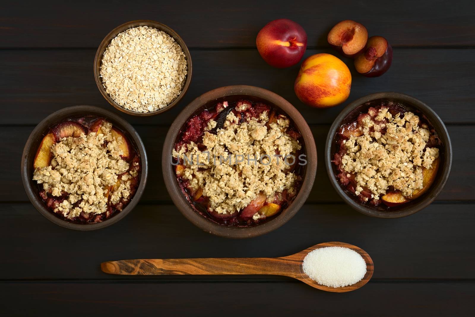 Overhead shot of three rustic bowls filled with baked plum and nectarine crumble or crisp, photographed on dark wood with natural light