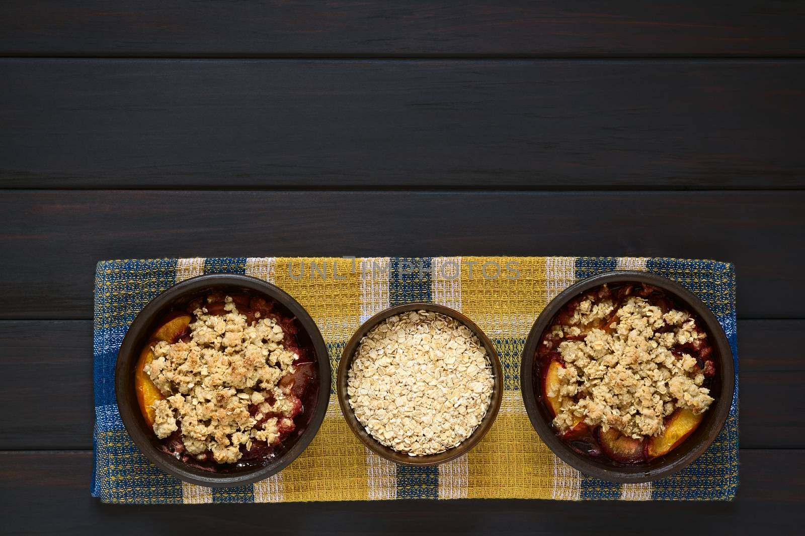 Overhead shot of two rustic bowls filled with baked plum and nectarine crumble or crisp and a bowl of rolled oats, photographed on cloth on dark wood with natural light