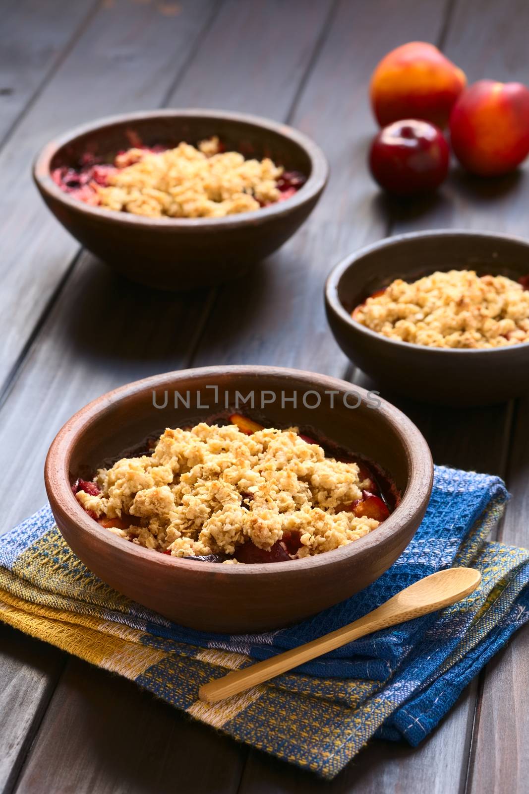 Three rustic bowls filled with baked plum and nectarine crumble or crisp, photographed on dark wood with natural light (Selective Focus, Focus one third into the first dessert)