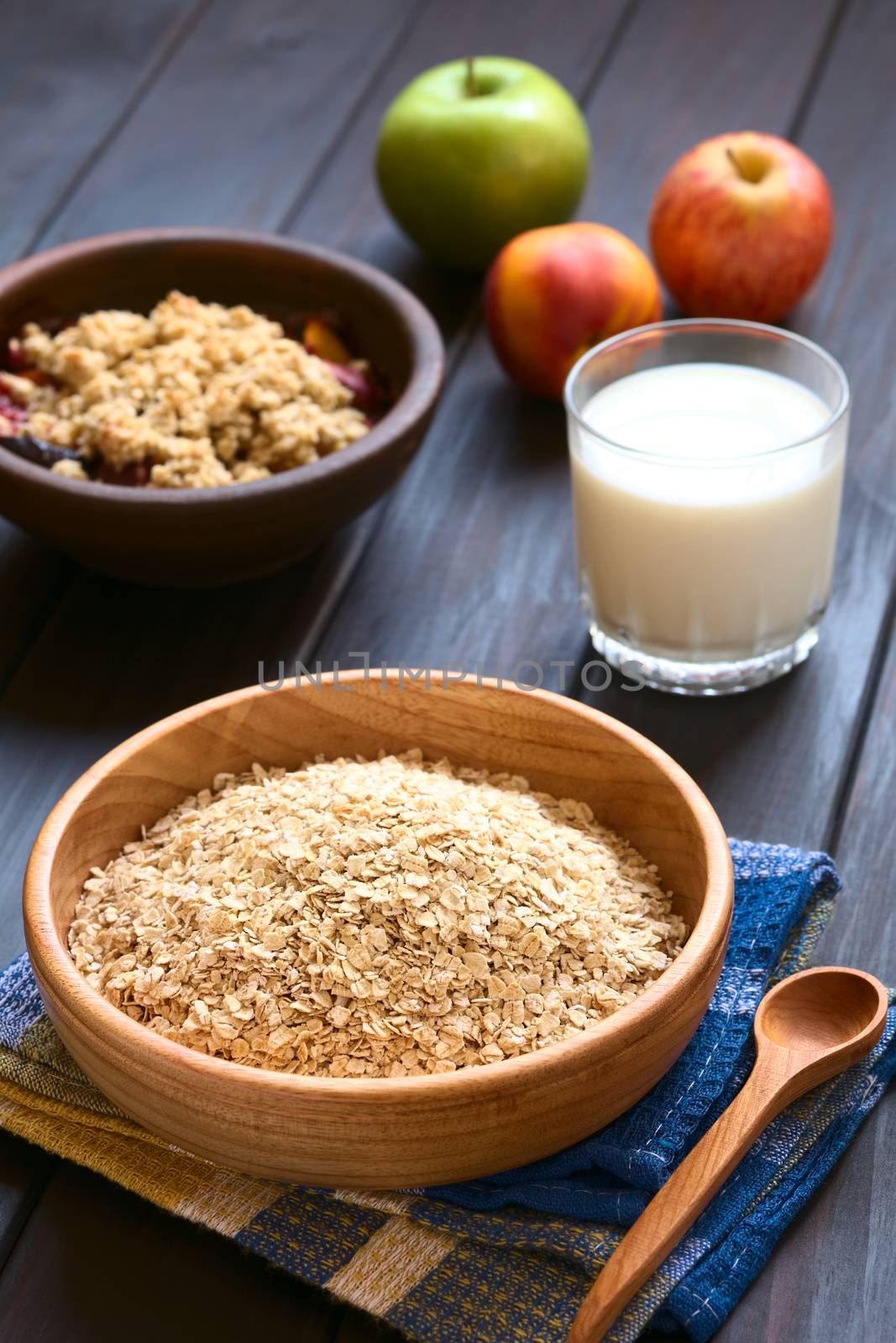 Raw rolled oats in wooden bowl with fruits, glass of milk and a bowl of fruit crumble in the back, photographed on dark wood with natural light (Selective Focus, Focus one third into the oats)