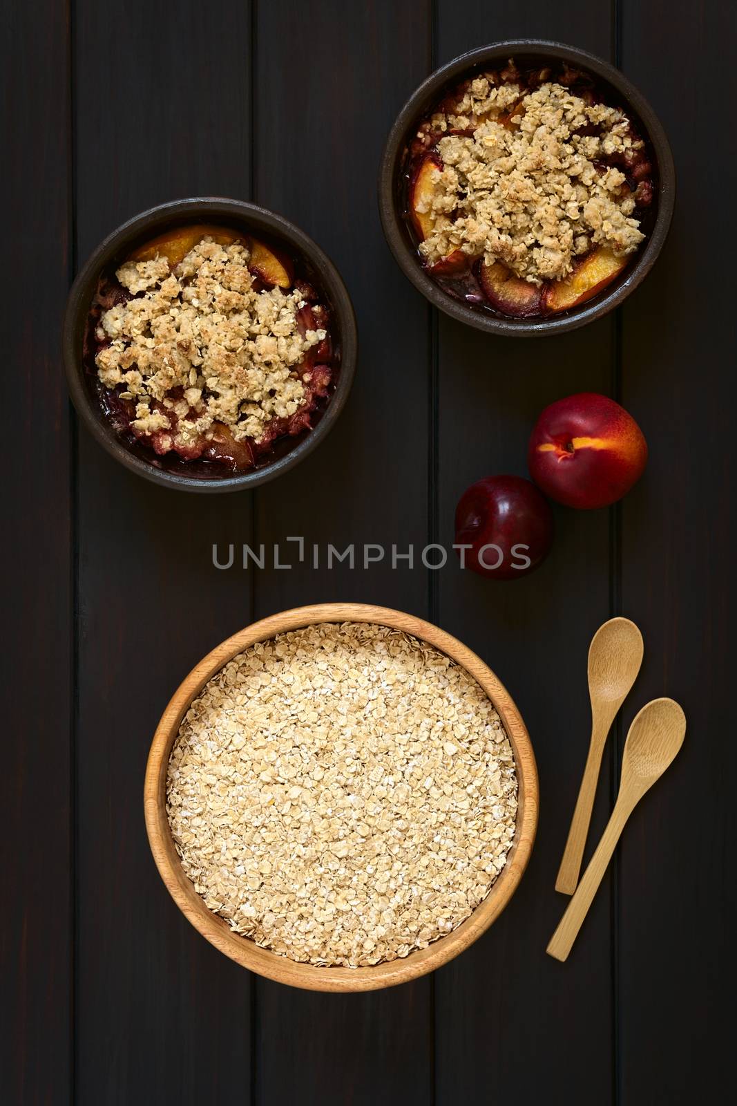 Overhead shot of a wooden bowl of rolled oats with two rustic bowls of plum and nectarine crumble, photographed on dark wood with natural light