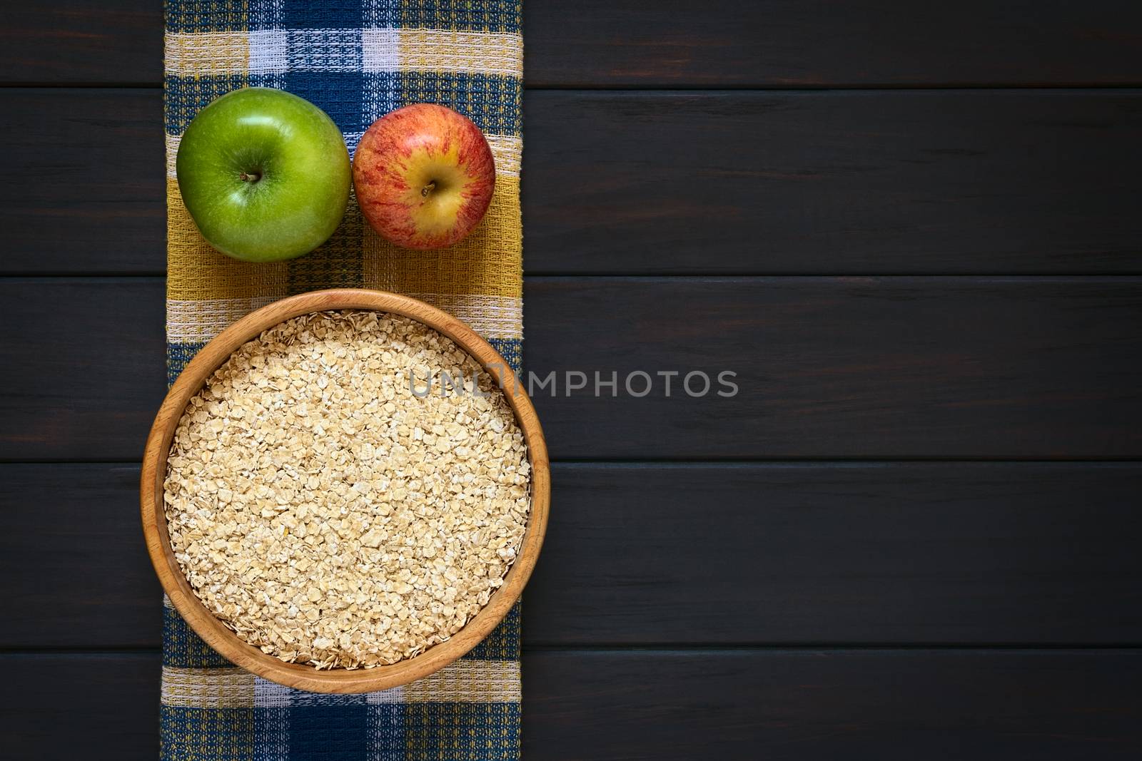 Overhead shot of raw rolled oats in wooden bowl with apples on cloth, photographed on dark wood with natural light