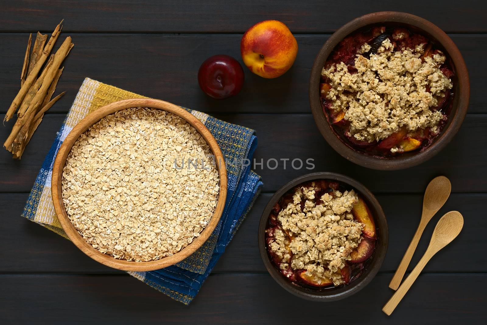 Overhead shot of a wooden bowl of rolled oats with two rustic bowls of plum and nectarine crumble, photographed on dark wood with natural light