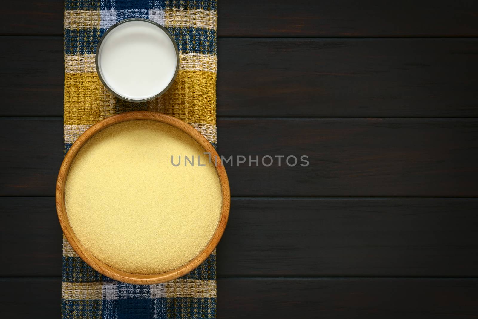 Overhead shot of raw semolina in wooden bowl with a glass of milk on cloth, photographed on dark wood with natural light  