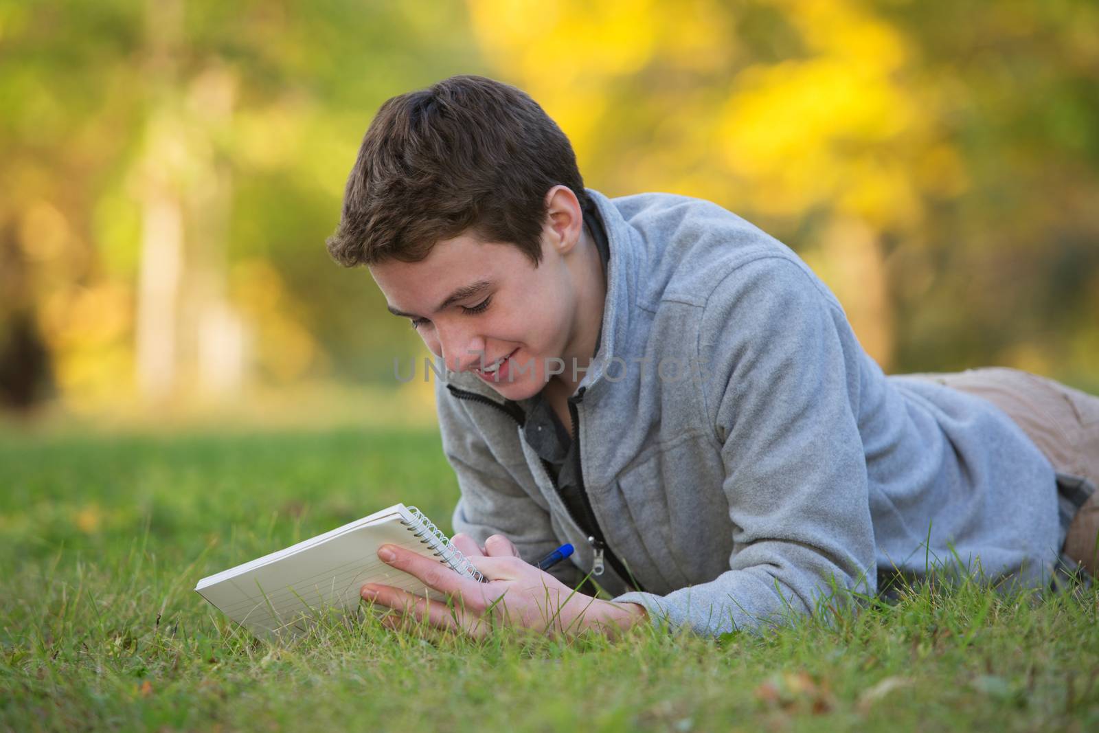 Smiling Caucasian teenager laying down on grass doing homework
