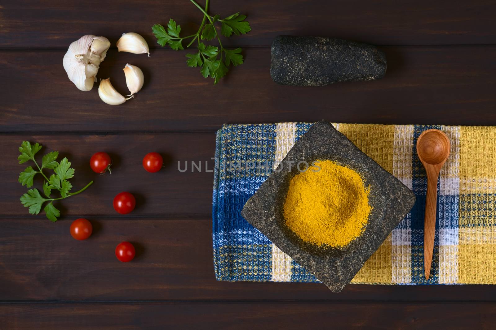 Overhead shot of curry powder spice in mortar with pestle, garlic, cherry tomatoes, parsley leaves on the side, photographed on dark wood with natural light 