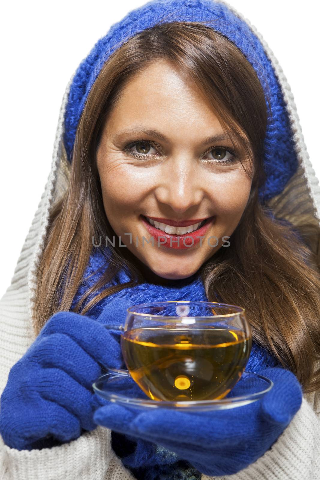 Fashionable young woman in a blue knitted winter ensemble and cowl neck jersey sipping a cup of hot tea with a smile in an effort to keep warm, isolated on white