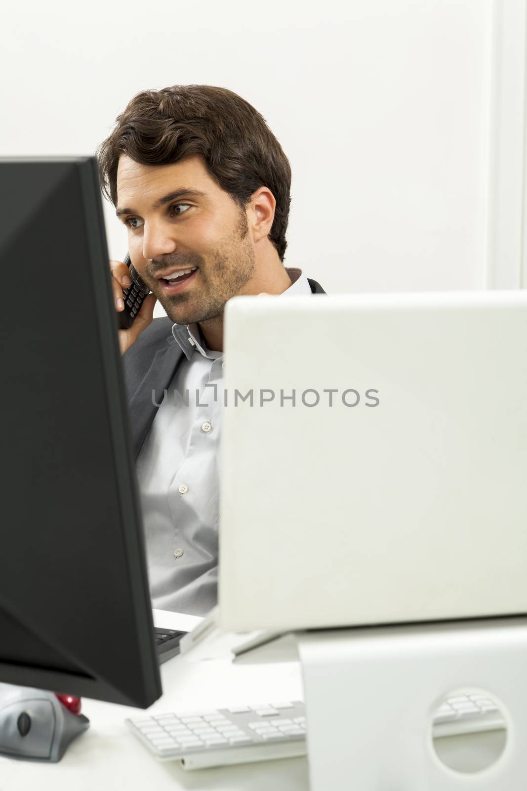 Successful businessman working in his office sitting in a chair at his desk and desktop computer listening to a call on his mobile phone, high angle side view