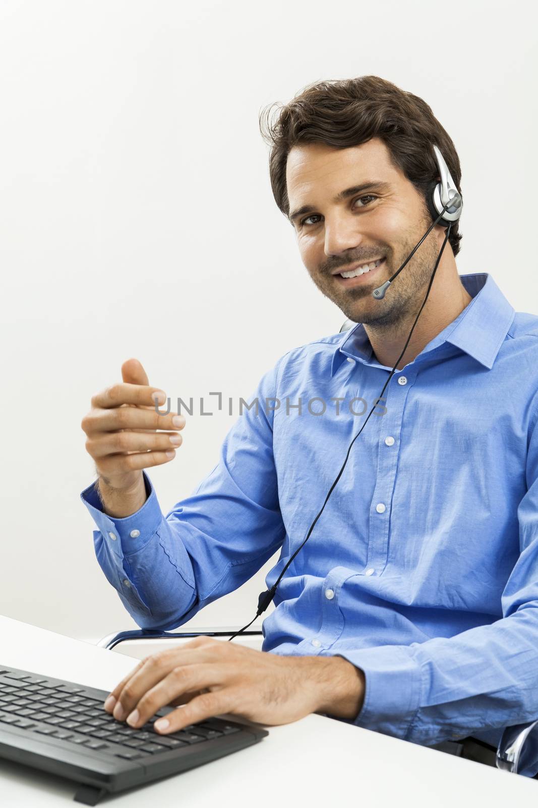 Attractive unshaven young man wearing a headset offering online chat and support on a client services of help desk as he types in information on his computer