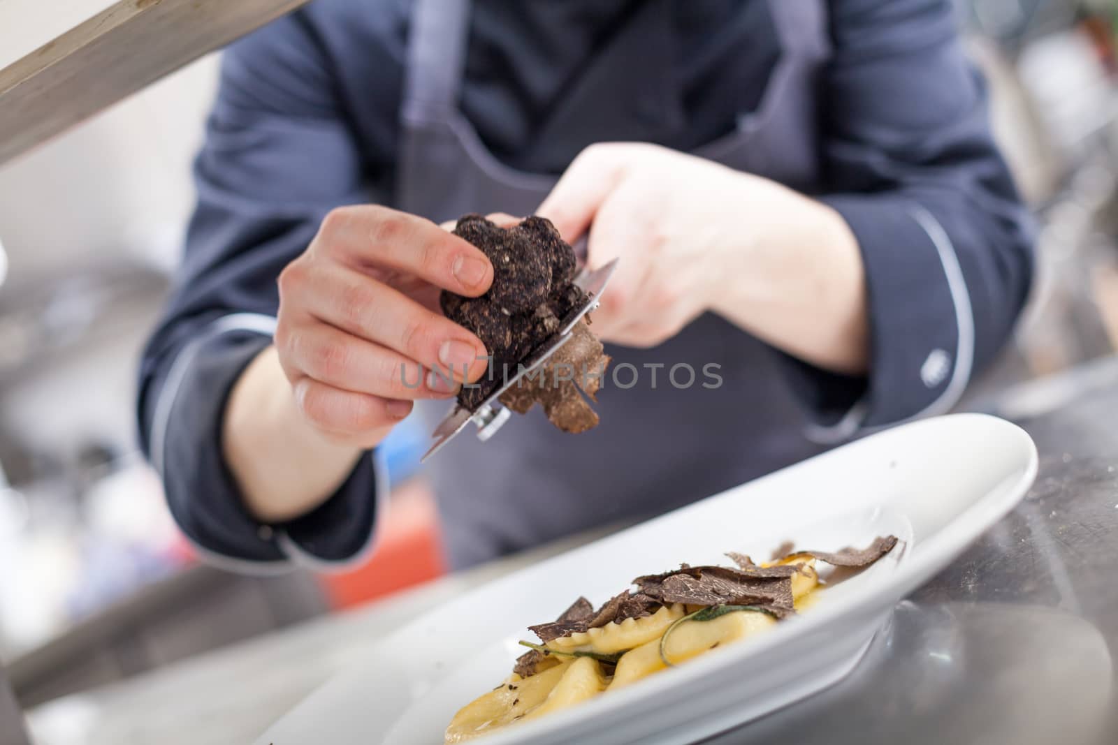 Chef grating truffle mushroom shavings onto homemade ravioli in a restaurant kitchen while preparing a dinner, close up view of the counter, plate and hands