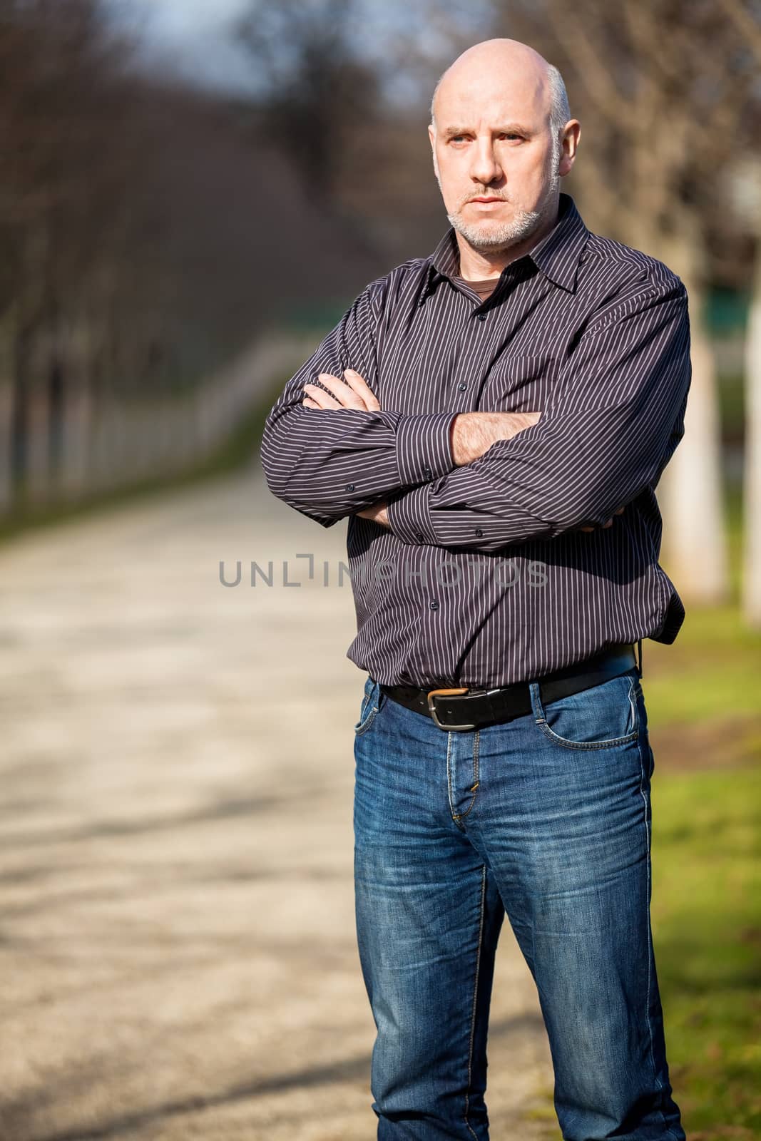 Confident attractive middle-aged man standing waiting in a rural lane with folded arms looking to the right hand side of the frame as though expecting somebody to arrive, with copyspace