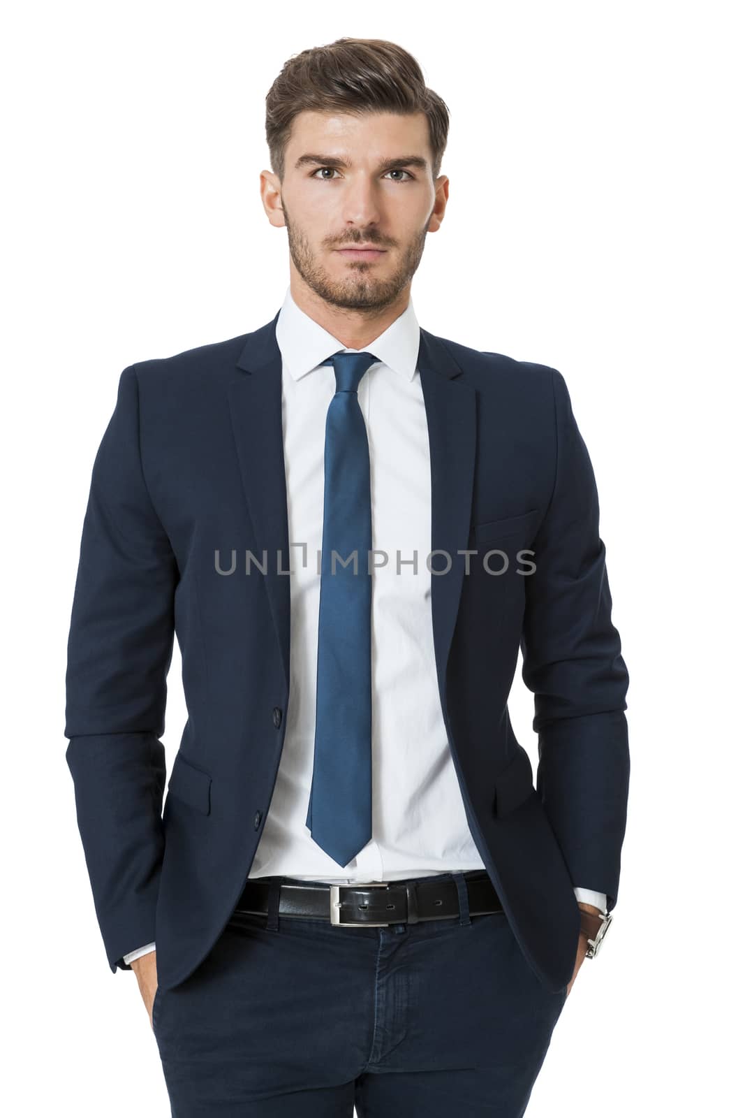 Stylish successful handsome young businessman standing in a relaxed pose with his hands in his pockets and his suit jacket open, isolated on white