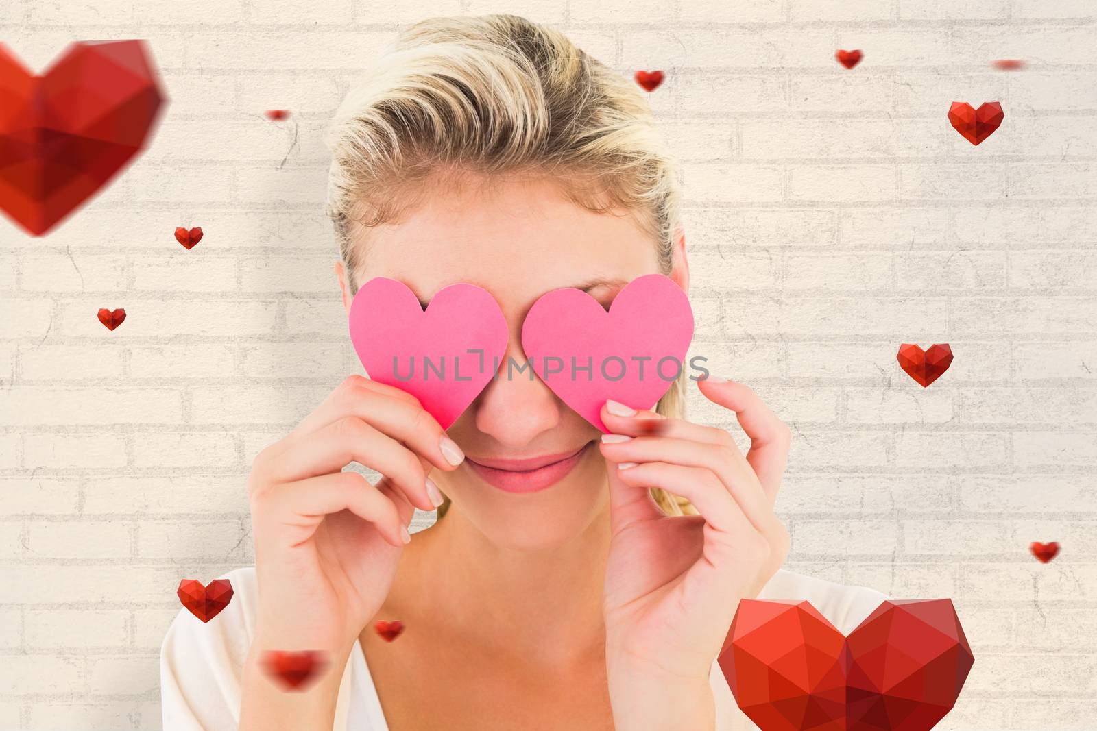 Attractive young blonde holding hearts over eyes against white wall