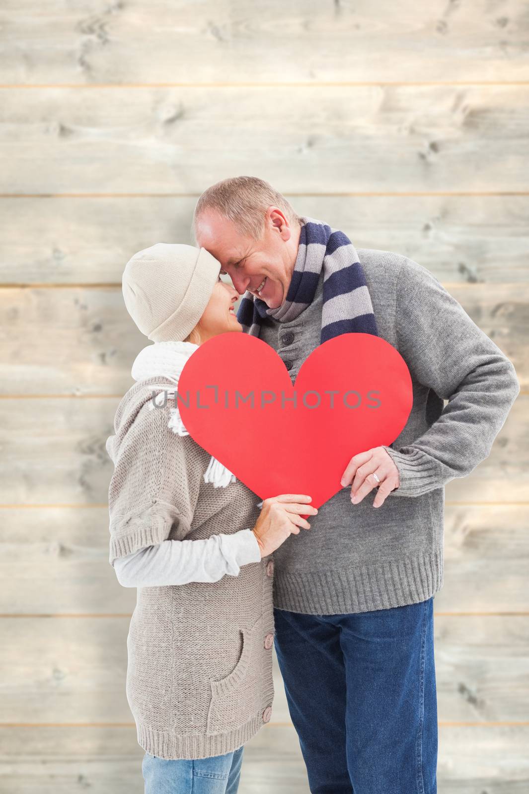 Happy mature couple in winter clothes holding red heart against pale wooden planks