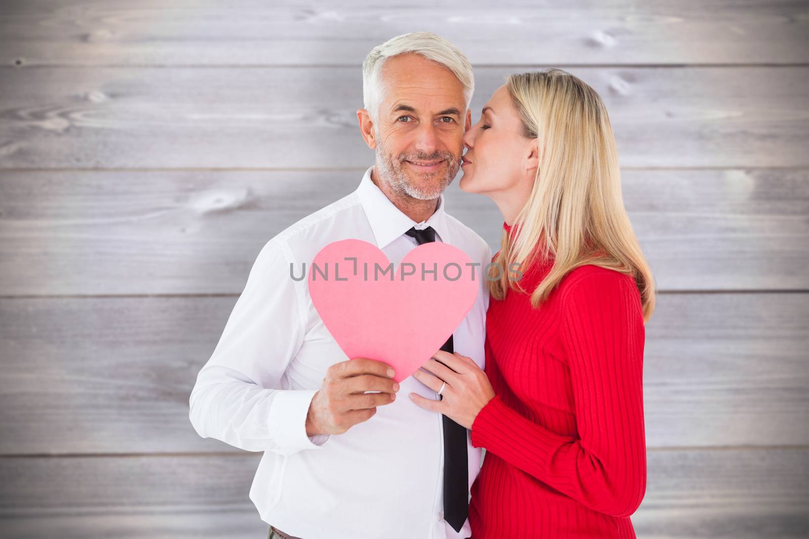 Handsome man holding paper heart getting a kiss from wife against wooden planks background