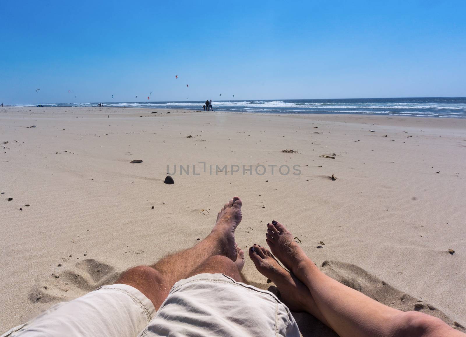 Two people relaxing on the beach with their feet in the sand watching wind surfers.