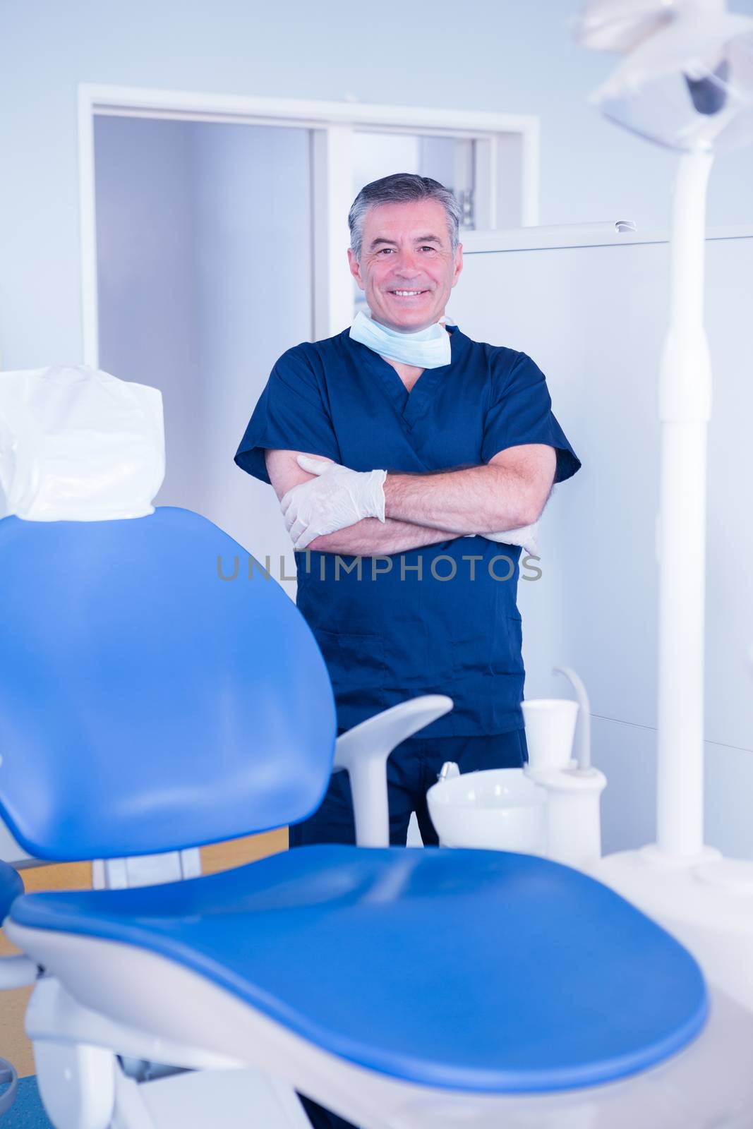 Dentist in blue scrubs smiling at camera beside chair by Wavebreakmedia