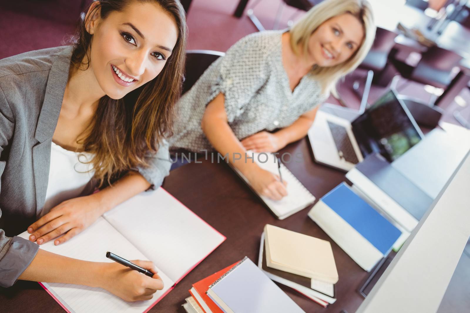 Smiling matures females students writing notes at desk in library