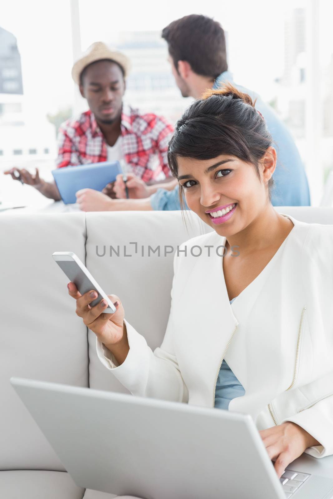 Smiling businesswoman using mobile and laptop on couch with colleagues behind her