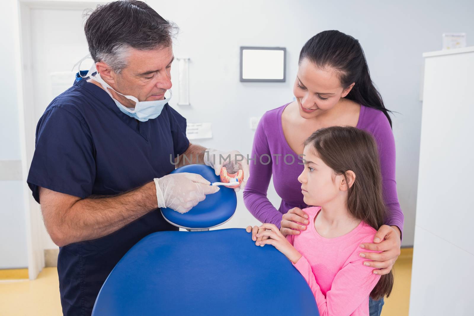 Dentist explaining to young patient and her mother how use toothbrush in dental clinic
