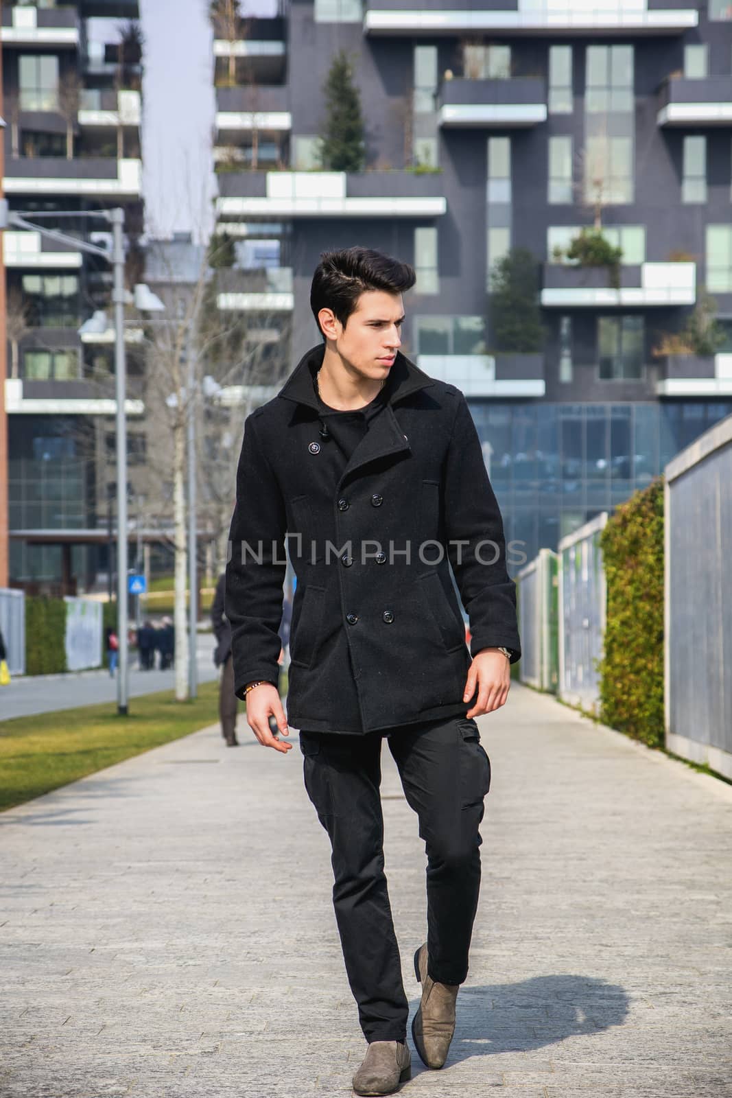 Stylish Young Handsome Man in Black Coat Standing in City Center Street with Skyscraper Behind Him, Looking to the Right
