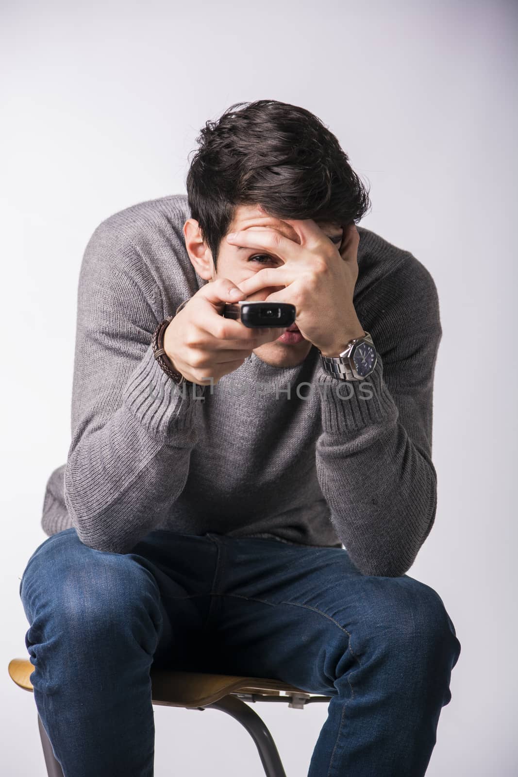 Handsome young man holding remote control and changing TV channels, covering his eyes scared sitting isolated on white