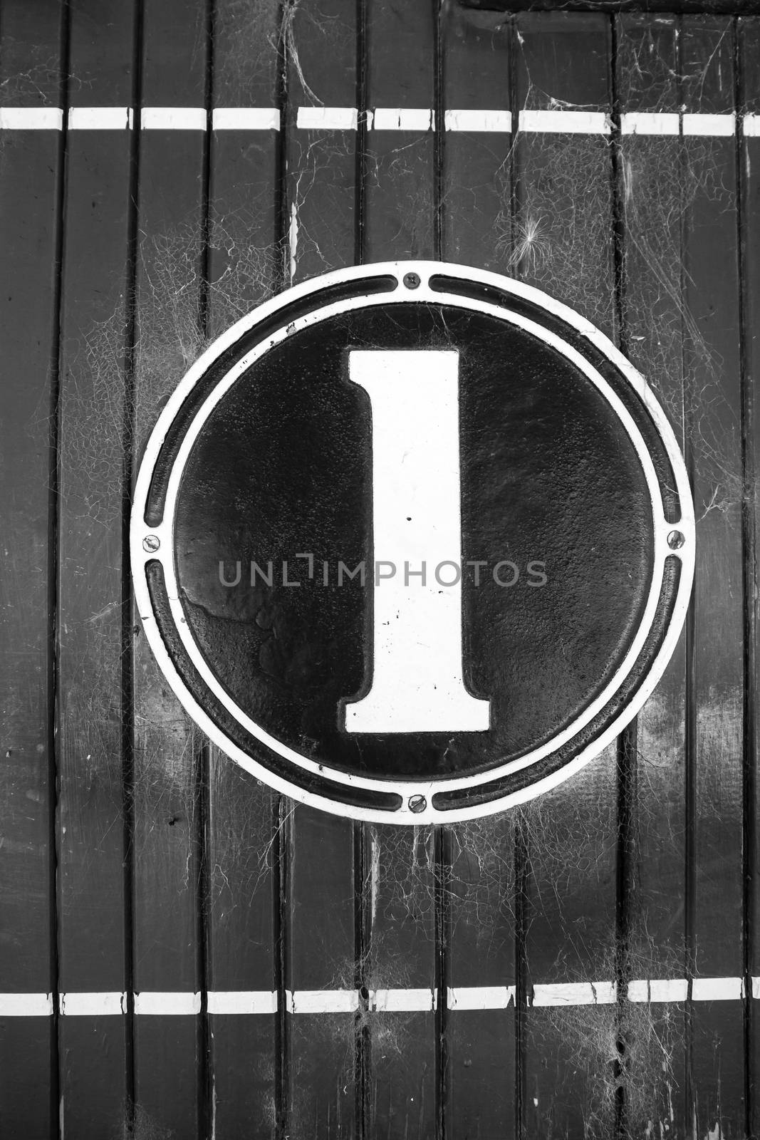 Numeral one, old fashioned sign, on circular cast metal and painted, mounted on wooden panelled wall in black and white retro style image. by brians101